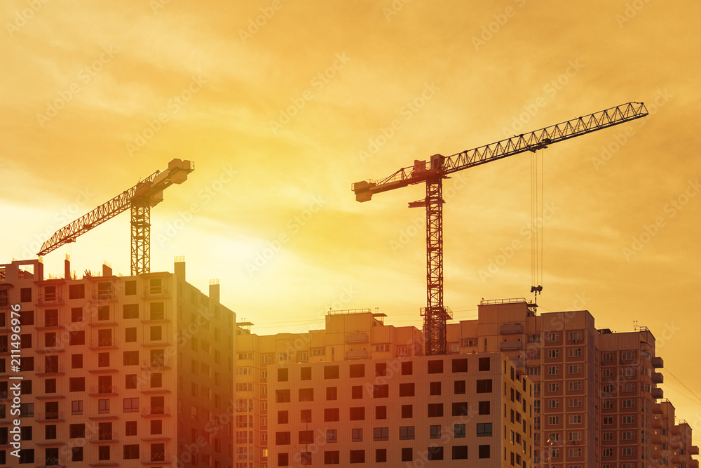 View of a dark silhouettes of two tower cranes building a modern panel houses against a orange sunset sky