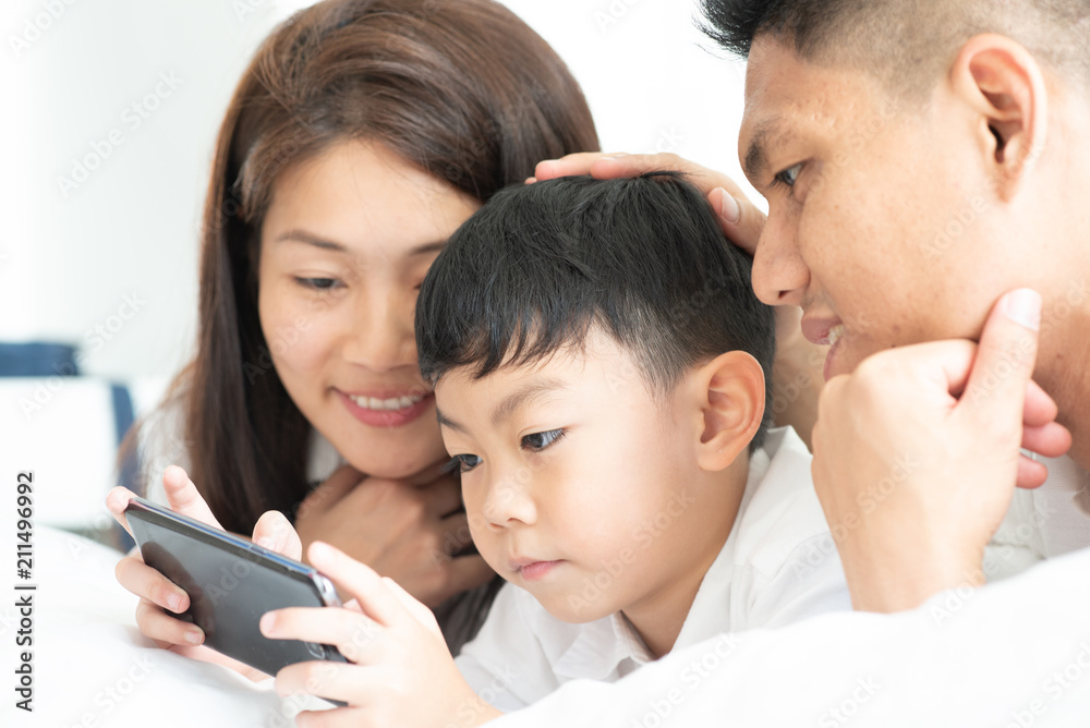 Asian boy playing smartphone with young parents lying in bed.Happy famility Asian boy playing smartphone with young parents lying in bed.Happy famility