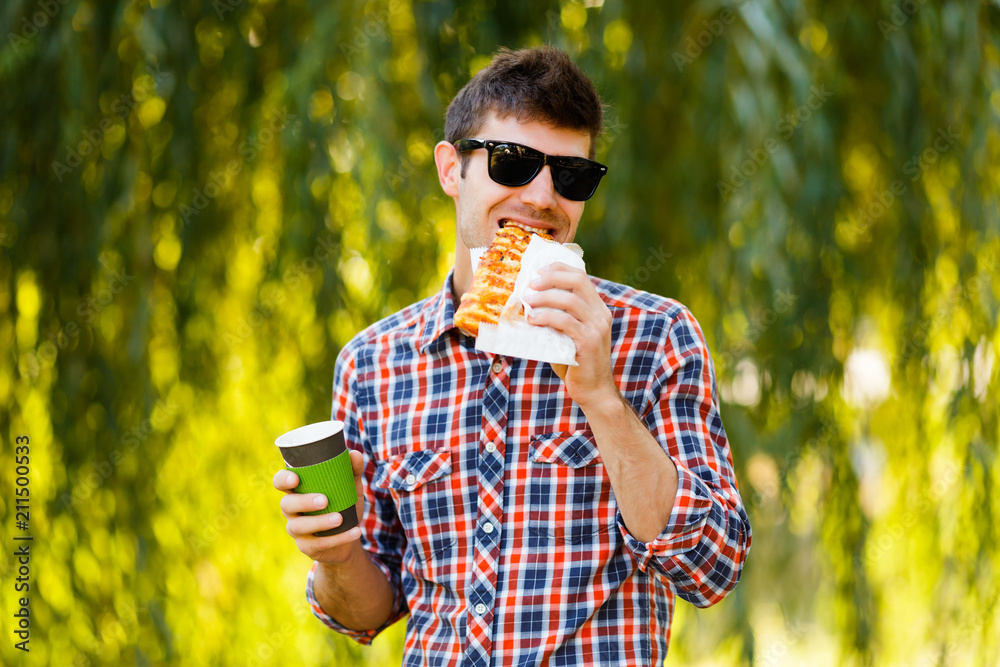 Fototapeta premium Man eating pizza. Cheerful young guy is eating pizza drinking coffee on the park