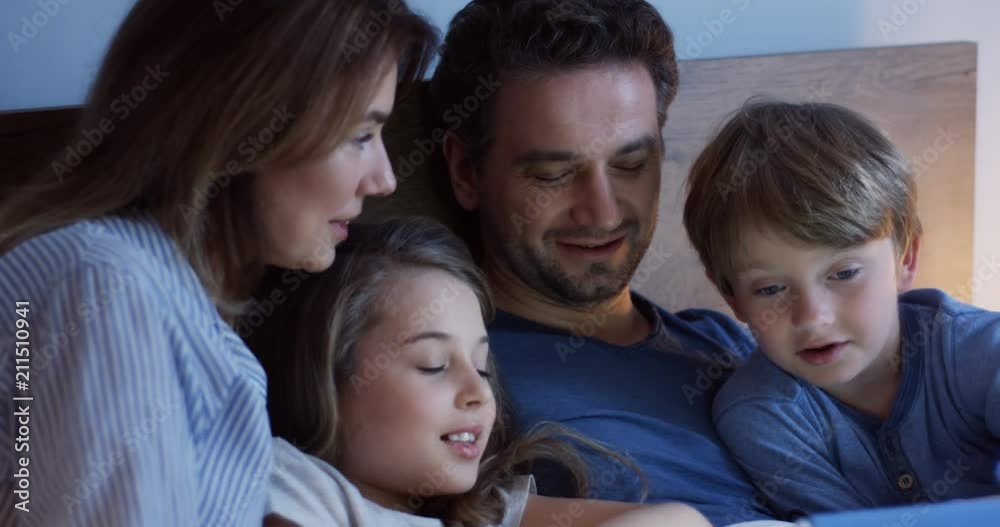 Close up of the smiling cute kids with their joyful parents lying in the bed late at night and watching video, cartoon or movie on the tablet computer. Indoor. Close up of the smiling cute kids with their joyful parents lying in the bed late at night and watching video, cartoon or movie on the tablet computer. Indoor.