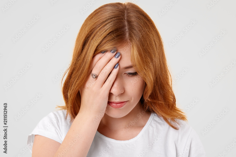 Portrait of serious female looks thoughtfully down, covers face with hand, feels lonely and bored, concentrated down, dressed in white casual t shirt, has manicure. Upset thoughtful woman indoor Portrait of serious female looks thoughtfully down, covers face with hand, feels lonely and bored, concentrated down, dressed in white casual t shirt, has manicure. Upset thoughtful woman indoor