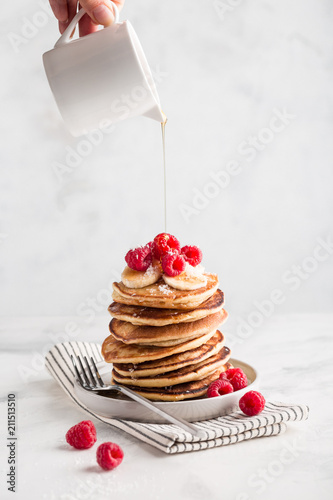 Hand pouring maple syrup on stack of homemade pancakes with fresh raspberries on light marble background