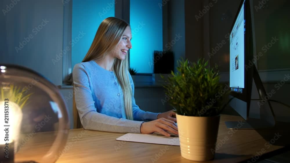 Smiling woman working on computer at night. Smiling female in checkered shirt sitting at lit by small lamp wooden desk and looking excitedly at computer monitor writing down information. Smiling woman working on computer at night. Smiling female in checkered shirt sitting at lit by small lamp wooden desk and looking excitedly at computer monitor writing down information.