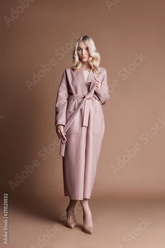 Beautiful blonde woman posing in a pink coat on a beige background. Fashion show clothing, woman with perfect figure, long hair. Trendy fashionable autumn coat on the girl's body