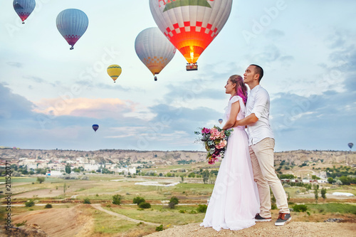 Date of a couple in love at sunset against background of balloons in Cappadocia, Turkey. Man and woman hugging standing on hill and look at big balloons. Engagement in the mountains of Cappadocia