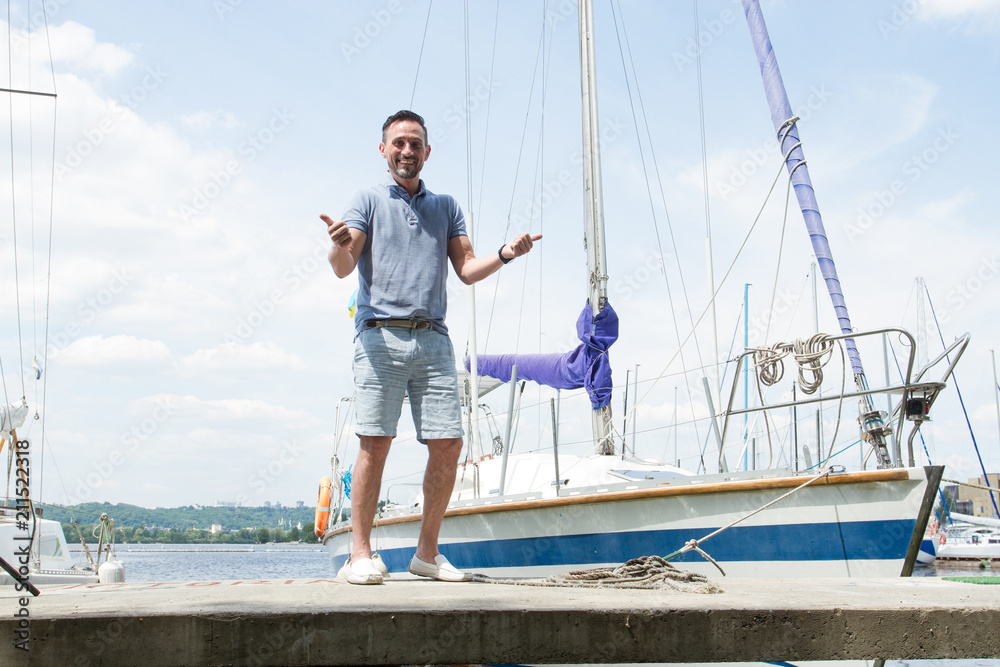 Smiling Yachtsman portrait on pier with both thumbs up. river and yachts on background. Young successful man sailor with own yacht. Man extremely have fun with yachting.