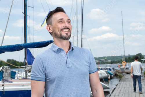 Young cheerful bearded man relaxing on sailboat posing and looking at far on background of boats, sky and folded wings. Portrait of yachtsman a fellow in blue polo sits on pier and boats on water.