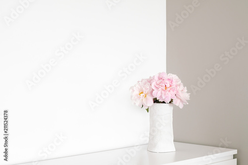Angle of white and gray walls in room. On dresser stands white vase with pink flowers blooming peonies.
