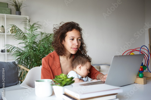 Motherhood, career and employment concept. Casually dressed young dark skinned single mother nursing baby and looking for job online, browsing websites, sitting at desk in front of open laptop