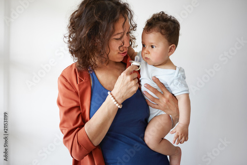 Картината върху платно Waist up shot of attractive caring young Hispanic mother nursing cute chubby baby boy, trying to soothe him to sleep, offering him pacifier