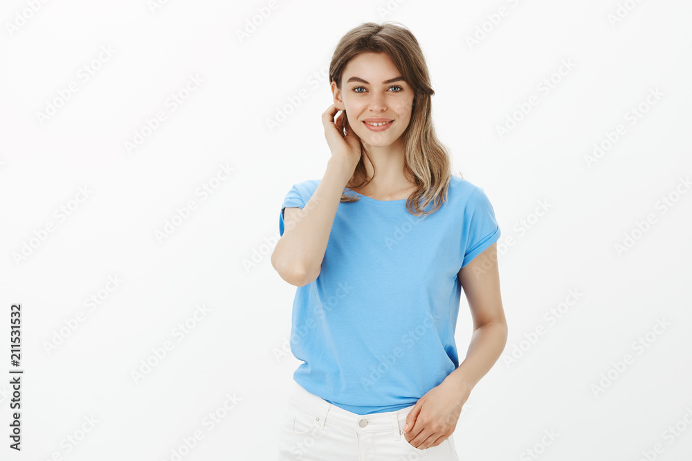 Portrait of positive charming fair-haired female in casual outfit, touching strand of hair and smiling broadly, being shy and tender, hearing compliment from cute guy over gray background Portrait of positive charming fair-haired female in casual outfit, touching strand of hair and smiling broadly, being shy and tender, hearing compliment from cute guy over gray background