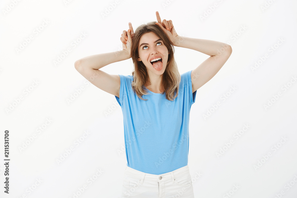 Fototapeta premium Girl being stubborn like goat feeling amused pissing off boyfriend with childish behavior. Positive good-looking woman, sticking out tongue, holding index fingers like horns and looking up