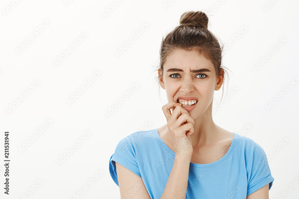 Waist-up shot of intense worried european woman with bun hairstyle, frowning, clenching teeth and biting fingernail anxiously, standing nervous over gray background, having terrible paranoia Waist-up shot of intense worried european woman with bun hairstyle, frowning, clenching teeth and biting fingernail anxiously, standing nervous over gray background, having terrible paranoia