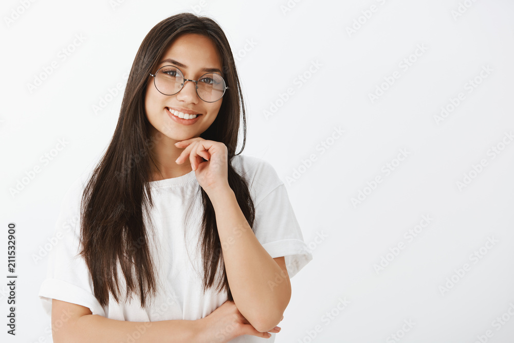 Waist-up shot of beautiful dreamy girlfriend in stylish eyewaer, holding hand near chin with hand crossed on chest, smiling broadly and tilting head while listening carefully with joy over gray wall Waist-up shot of beautiful dreamy girlfriend in stylish eyewaer, holding hand near chin with hand crossed on chest, smiling broadly and tilting head while listening carefully with joy over gray wall