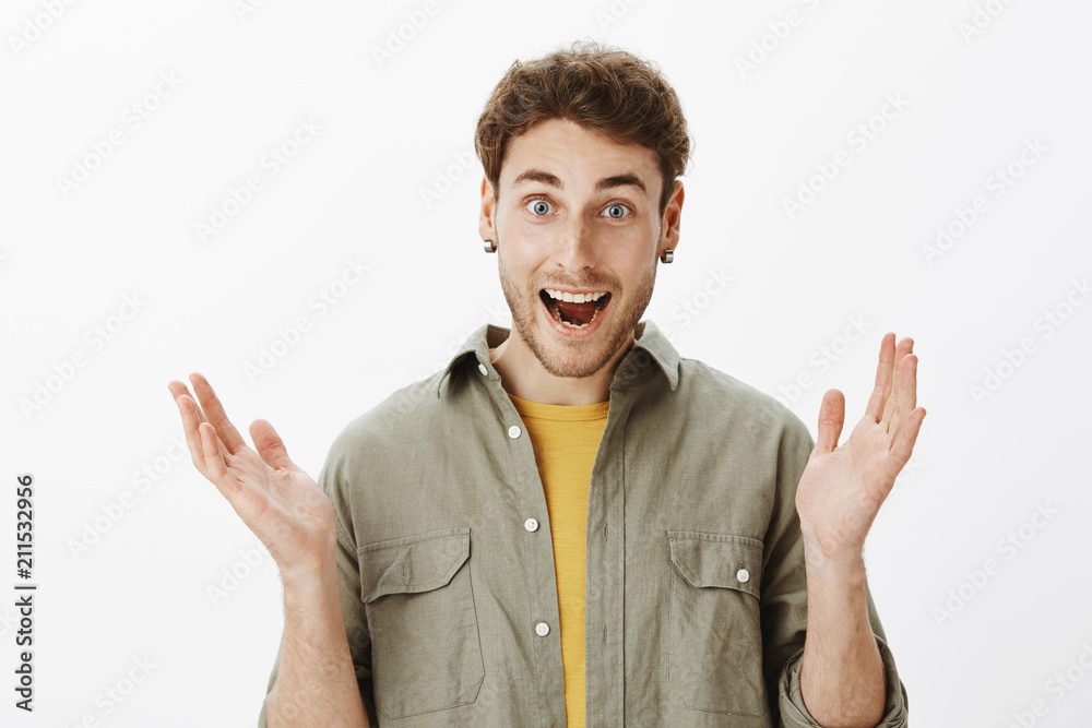 Fototapeta premium Amused good-looking happy male with beard and short curly hair, clasping in hands liking perfect perfomance of favorite band, smiling broadly and gazing with delight at camera, posing over gray wall