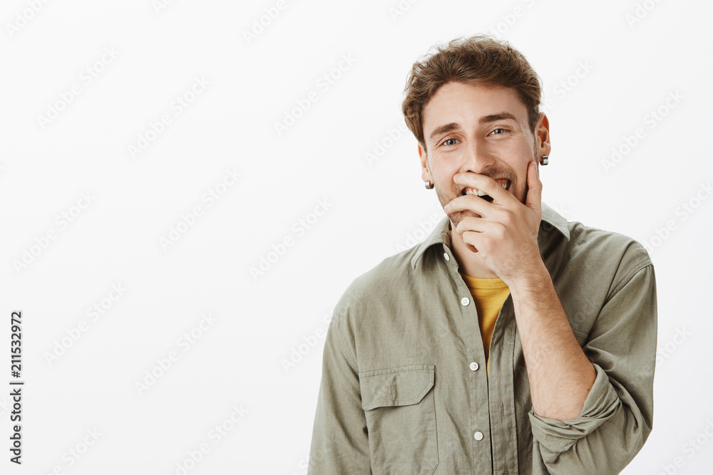 Indoor shot of funny handsome man with short curly hair and beard, covering opened mouth while smiling and laughing, standing amused and joyful over gray background, enjoying great time with friends Indoor shot of funny handsome man with short curly hair and beard, covering opened mouth while smiling and laughing, standing amused and joyful over gray background, enjoying great time with friends