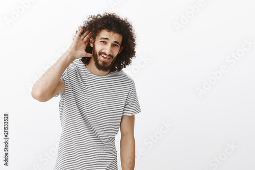 Studio shot of carefree good-looking mature guy with beard and afro hairstyle  holding hand near ear and asking question  being at concert with friend  hearing nothing at loud party over gray wall