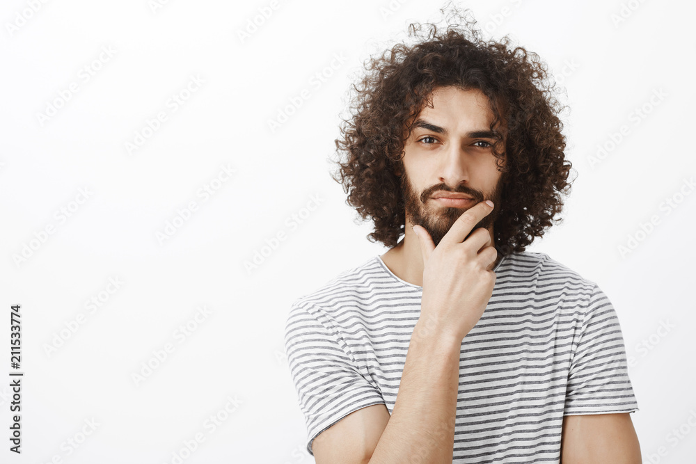 Smart thougtful attractive hispanic guy with beard and curly hair, holding hand on chin, thinking and focusing at camera while making decision or picking something in store, standing over gray wall Smart thougtful attractive hispanic guy with beard and curly hair, holding hand on chin, thinking and focusing at camera while making decision or picking something in store, standing over gray wall
