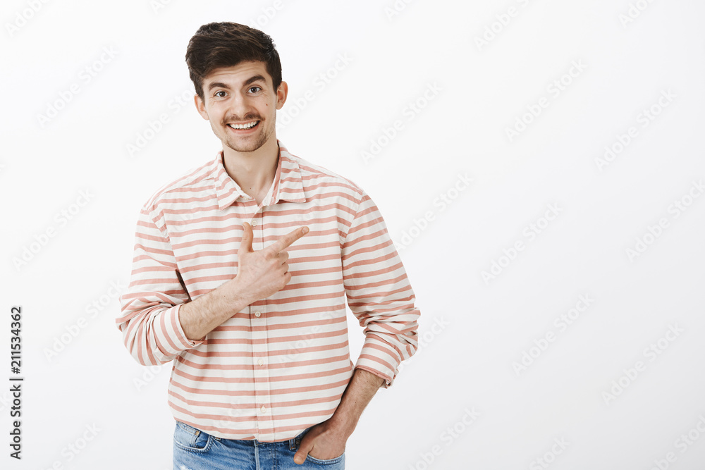 Indoor shot of friendly funny caucasian man with moustache, bending towards camera and laughing while pointing at upper right corner, discussing hilarious advertisement with broad smile over gray wall Indoor shot of friendly funny caucasian man with moustache, bending towards camera and laughing while pointing at upper right corner, discussing hilarious advertisement with broad smile over gray wall