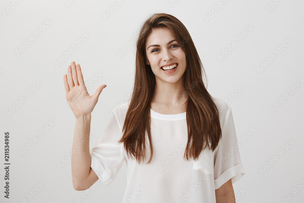 Friendly woman says hi to new neighbours. Portrait of charming young emotive woman waving with raised hand, greeting or welcoming close friend, smiling broadly at camera, standing over gray wall Friendly woman says hi to new neighbours. Portrait of charming young emotive woman waving with raised hand, greeting or welcoming close friend, smiling broadly at camera, standing over gray wall