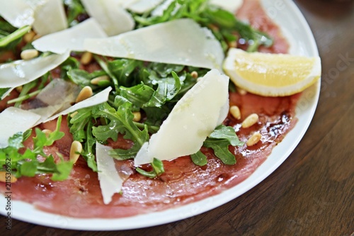 Delicious and healthy Raw Food: Beef carpaccio on a white round plate with parmesan cheese, rocket salad and lemon, drizzled with extra virgin olive oil by a professional Starred Chef. Natural light.