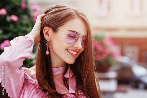 Outdoor close up portrait of young beautiful happy smiling woman wearing pink aviator sunglasses, trendy earrings, posing in street of european city. Copy, empty space for text