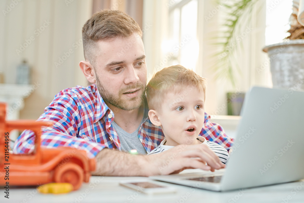 Warm toned portrait of handsome father teaching son how to use laptop or playing computer games together, focus on excited little boy