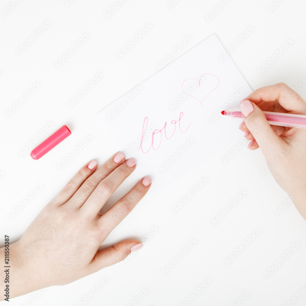 Fototapeta premium Flat lay. Top view. Hand of girl writing love letter on Saint Valentines Day. Handmade postcard with red heart shaped figure. Woman write Love on postcard for 14 February holiday celebration