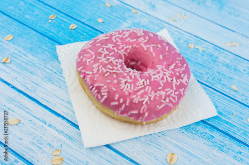 Tasty Sweet Pink Donut On Paper On Scratched Blue White High Contrast Wood Background With Whole Grains Oat Meal On It