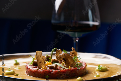Beef tartare Steak with egg yolk, pistachios, truffle oil, green mayonnaise on porcelain plate and glass of red wine on restaurant table. Classic tartare meat with ingredients. Luxury food