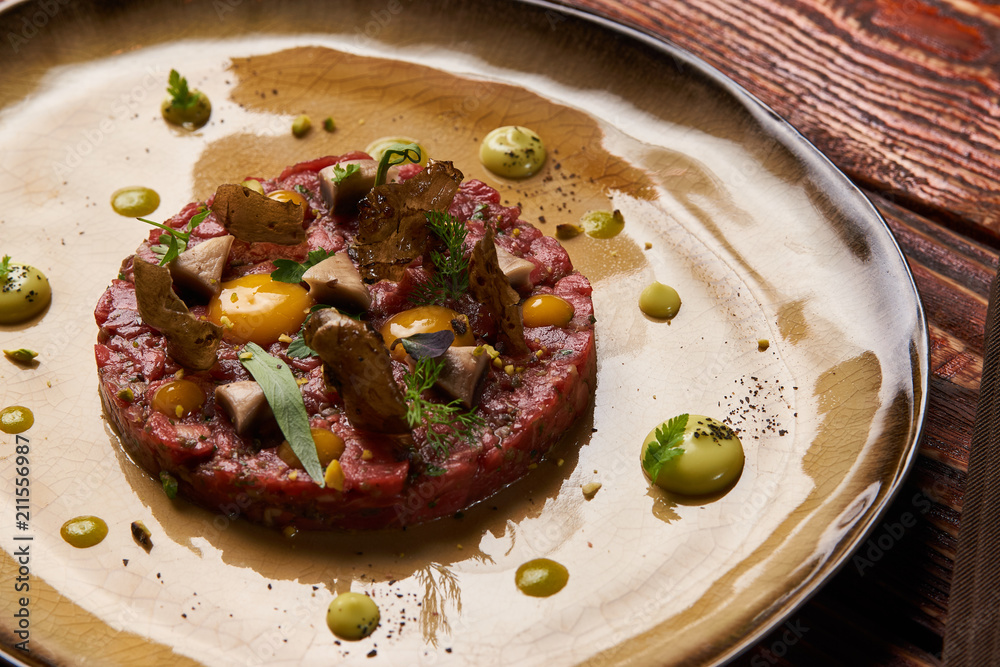 Tartare Steak with egg yolk, pistachios, truffle oil, green mayonnaise on plate and glass of red wine, served on restaurant table. Classic Beef tartare meat, Luxury food