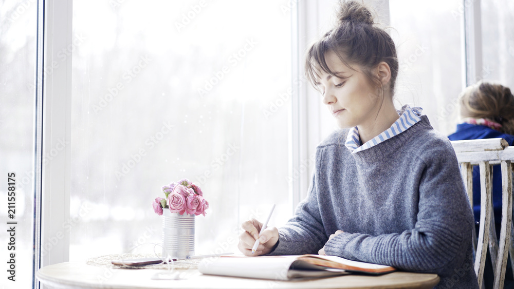 A young brunette girl in a cafe sitting near the window dressed in a blue pullover is reading a notebook in the cafe holding a pencil.