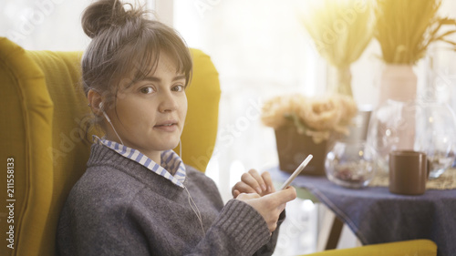 A portrait profile of a young white cute girl in earphones listening to music holding a white phone sitting in a big yellow chair looking at the camera