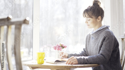 A smiling calm young brunette girl in a cafe sitting near the window dressed in a blue pullover reading a notebook holding a pencil