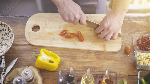 A closeup of male hands cutting slicing cherry tomatoes on a wooden kitchen cooking board olive and ingredients surrounding