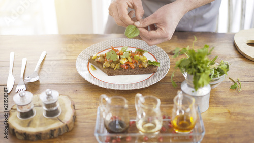 A male chief is adding pieces of parsley to a stew salad on a piece of a black bread