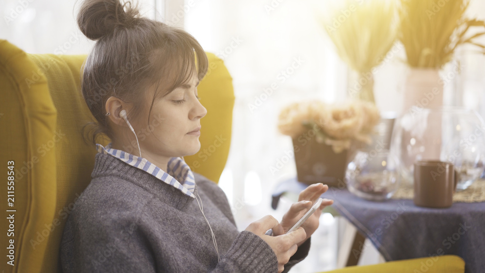 A portrait profile of a young white cute girl in earphones listening to music holding a white phone sitting in a big yellow chair
