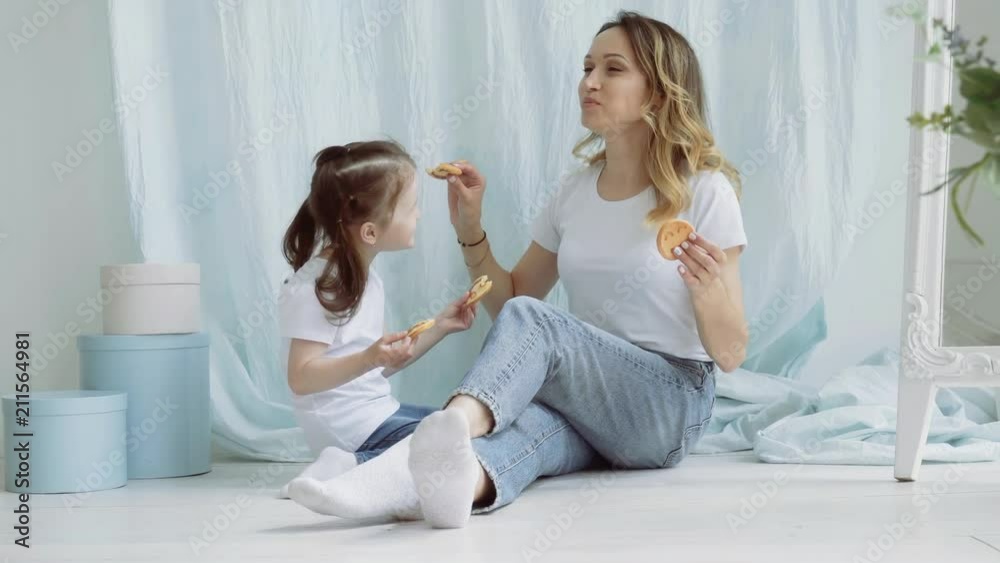 Mom and little girl feed each other cookies. A woman with a little daughter are sitting on the floor. Mom and little girl feed each other cookies. A woman with a little daughter are sitting on the floor.