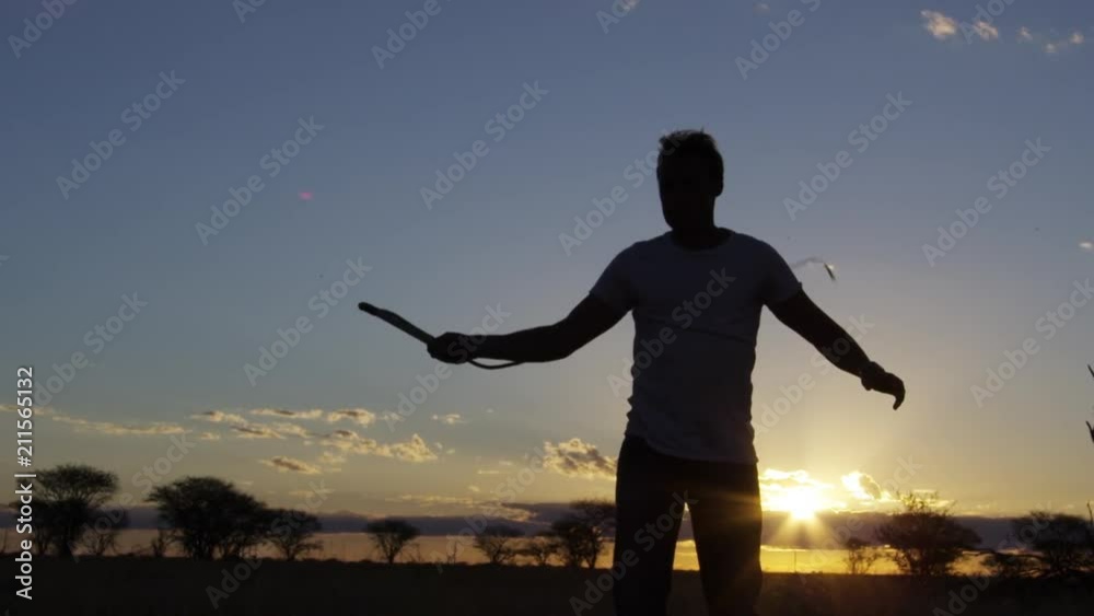 A medium shot of a silhouetted man whipping his whip out on a field with trees in slow motion. A medium shot of a silhouetted man whipping his whip out on a field with trees in slow motion.