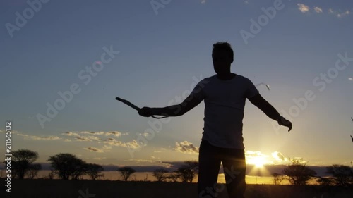 A medium shot of a silhouetted man whipping his whip out on a field with trees in slow motion.