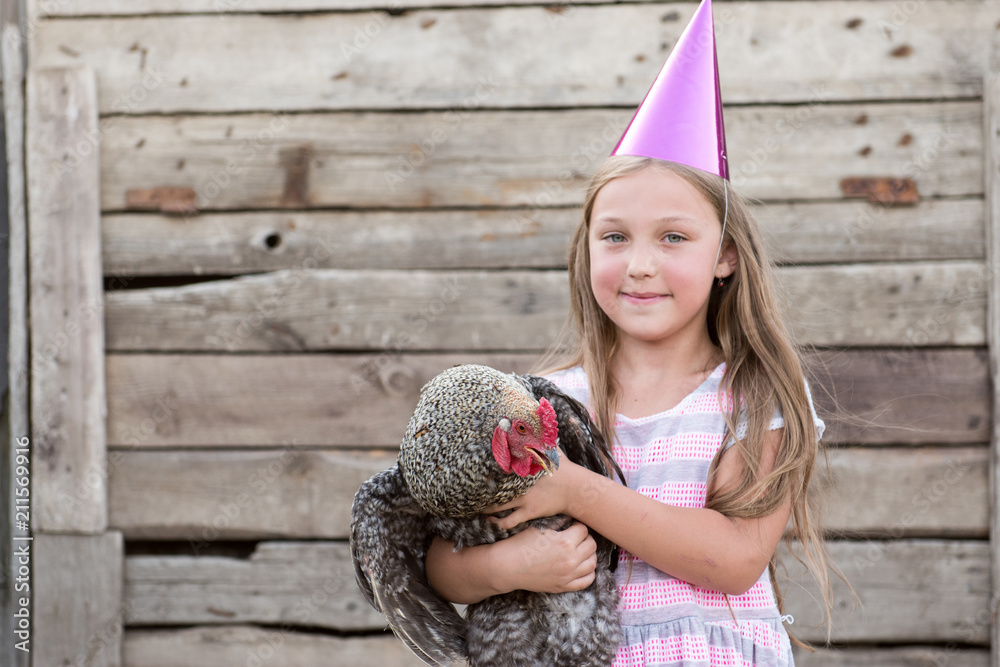 Young girl in pointed magic hat holds motley chicken in her hands and poses in front of camera in village courtyard. The concept of healthy lifestyle in nature, love of the world, respect for nature