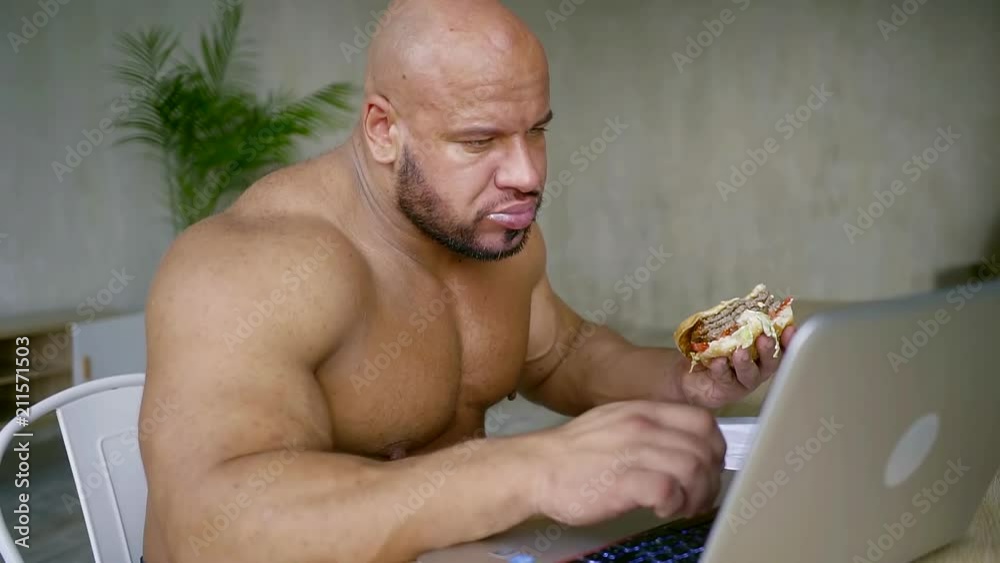 naked strong man is eating beefburger, sitting at table with notebook and typing on it, lunch in home