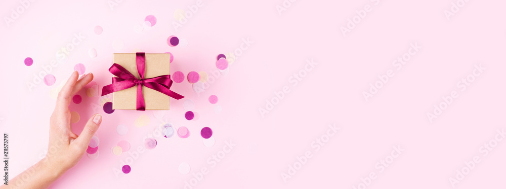 Woman hand and kraft gift box with pink bow on pink background decorated with confetti.. Top view, holiday present concept. Banner for site.