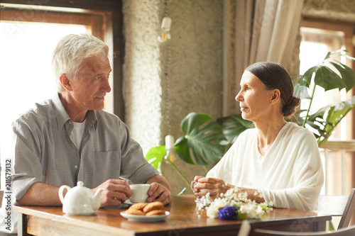 Restful senior couple sitting by table in cafe  having tea with cookies and talking