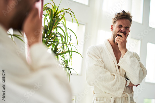 Waist up portrait of drowsy man yawning after night. He is standing in front of mirror in bathrobe and covering face with hand