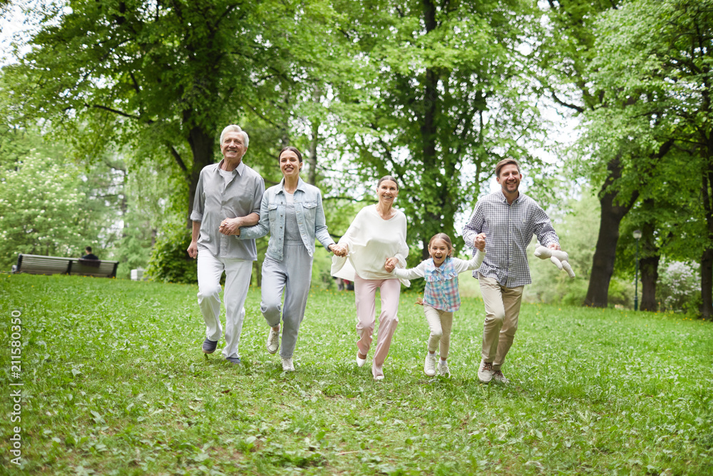 Obraz premium Cheerful and energetic family running on green grass among trees in summer park