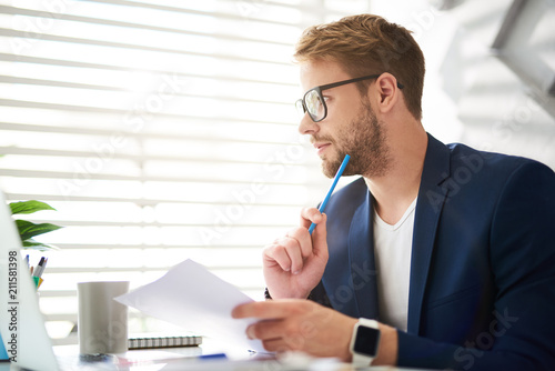 Profile of handsome young male sitting at table and holding papers and blue pencil in hands. He is looking sideways absorbed in personal consideration of business ideas