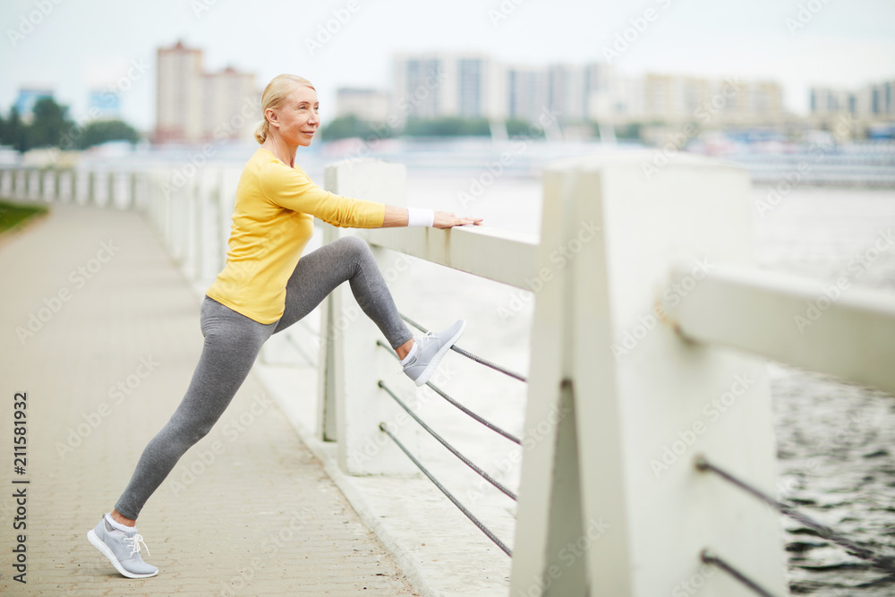 Obraz premium Mature blonde in activewear standing by riverside on city bridge and doing exercise for legs