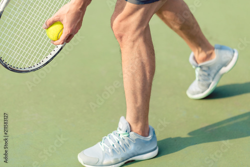 Low section of a professional player wearing gray sport shoes while holding the ball and the tennis racket during match on green surface
