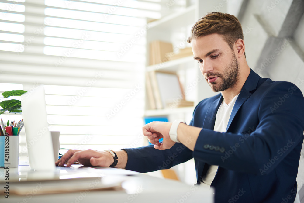 Have to go. Side view of occupied businessman sitting at table and looking at watch. He is working in office with concentration